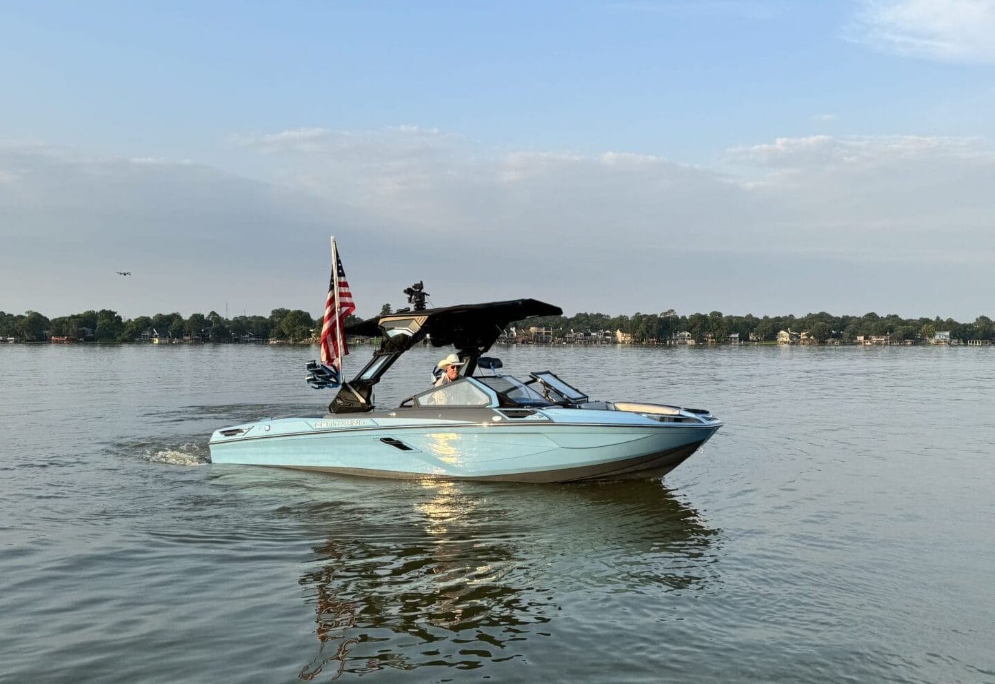 A light blue motorboat with an American flag moves across a calm lake under a partly cloudy sky, with a tree-lined shore in the background.