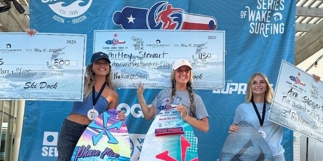 Three women stand on a podium holding wakeboards and oversized prize checks at a wake surfing competition, with event banners in the background.