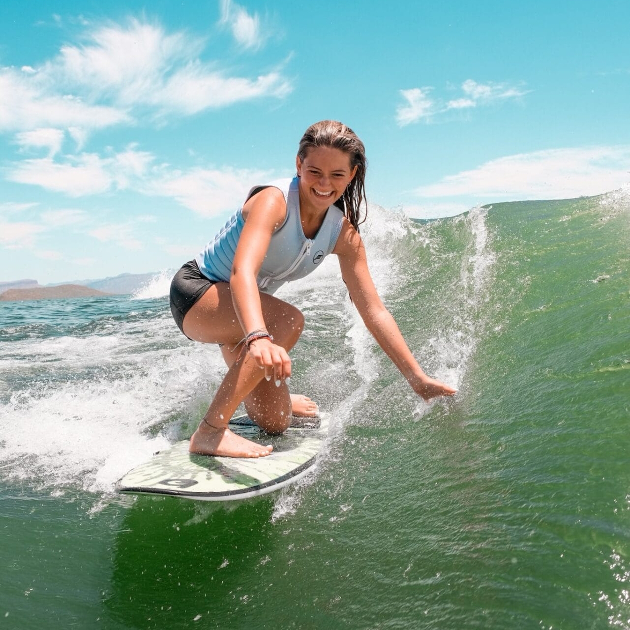 A woman in a white sleeveless top and black shorts rides a wave on a surfboard under a clear blue sky.
