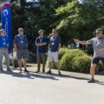 A group of men stand outdoors watching as one man in athletic wear gestures forward, appearing to participate in an activity or game.