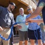 Four adults stand outdoors, three holding a large knotted blue rope while one person hands a paper to a woman in sunglasses.