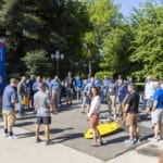 A group of people stands in a circle outdoors on a sunny day, listening to a speaker near a yellow kayak and a vertical event banner. Trees and greenery are in the background.