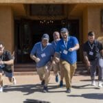 A group of men wearing name badges start a race outside a building, smiling and appearing energetic in the sunlight.