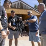 Four men stand outside a wooden building, closely examining a mechanical device or robot that one of them is holding.