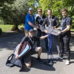 Five men pose outdoors on a sunny day with electronic gear, cables, and a surfboard, standing and kneeling on a paved path surrounded by greenery.