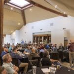 A man stands speaking to a seated audience in a large, well-lit room with wooden beams and a skylight.