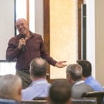 A man stands at the front of a room speaking into a microphone and gesturing with one hand, while several people seated in chairs listen.