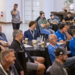 A group of people sit at round tables in a conference room, listening attentively to a presentation. Some have drinks and notepads in front of them.