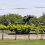 A black motorboat on a trailer is being towed down a road, partially obscured by green vineyard plants in the foreground.