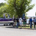 A group of people stands near a purple and white boat on a trailer, parked by a curb under trees on a sunny day.