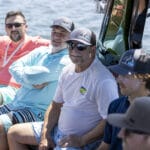 Five men sit together on a boat in casual summer clothing, wearing hats and sunglasses, with water in the background.