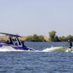 A person is wakeboarding on a lake, being towed by a purple motorboat with several people onboard. Trees and grassy land are visible in the background.
