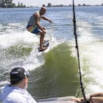 A man in a life vest surfs on a wave behind a boat, while three people in the boat watch him.