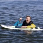 A man wearing a life vest floats in the water with a paddleboard, smiling and giving a thumbs-up gesture.