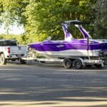 A white pickup truck towing a purple and white speedboat on a trailer is parked on a paved road surrounded by green trees and bushes.