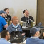 A group of men seated at round tables listen attentively and smile during a conference or meeting.