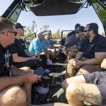 A group of men sit and talk inside a boat on a sunny day, some holding their phones and water bottles, with trees visible in the background.