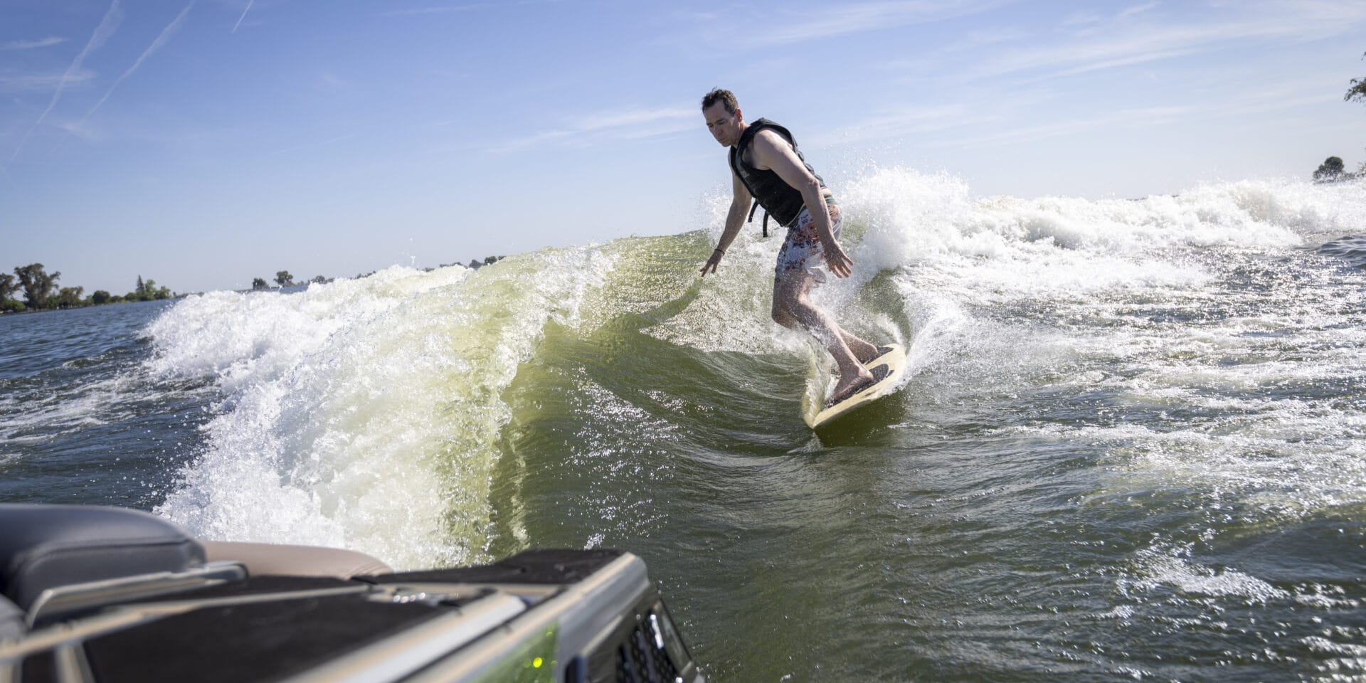 A person wearing a life vest is wake surfing on a wave behind a green boat on a sunny day.