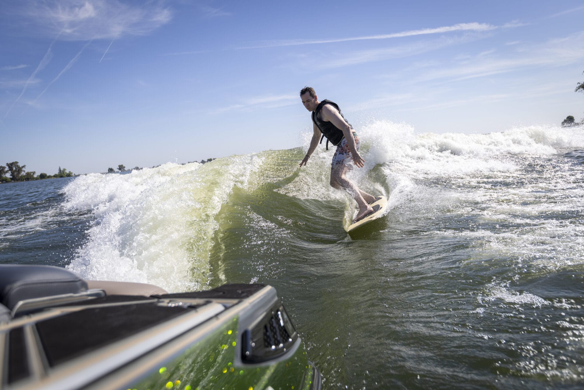 A person wearing a life vest is wake surfing on a wave behind a green boat on a sunny day.
