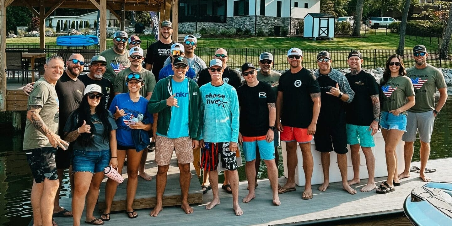 A group of people pose for a photo on a dock by the water, with trees, a gazebo, and a house visible in the background.
