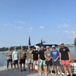 A group of ten men stands barefoot on a dock in front of a boat, with a lake and trees visible in the background on a sunny day.