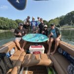 Eight people pose on a boat with a wakeboard, wearing casual clothes and hats, with trees and water visible in the background.