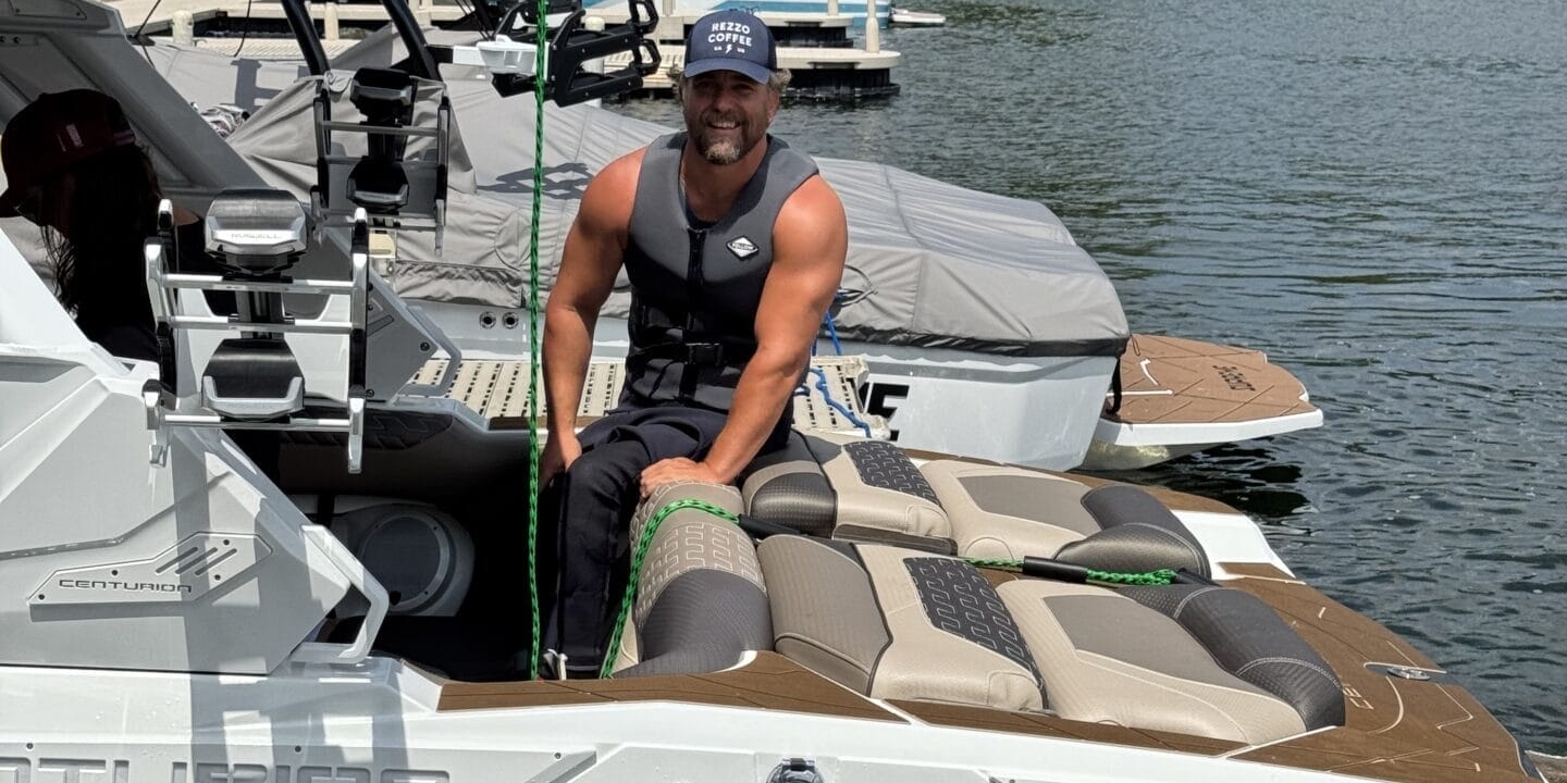 A person wearing a life jacket and cap sits on the edge of a boat docked on a lake, surrounded by trees and other boats.