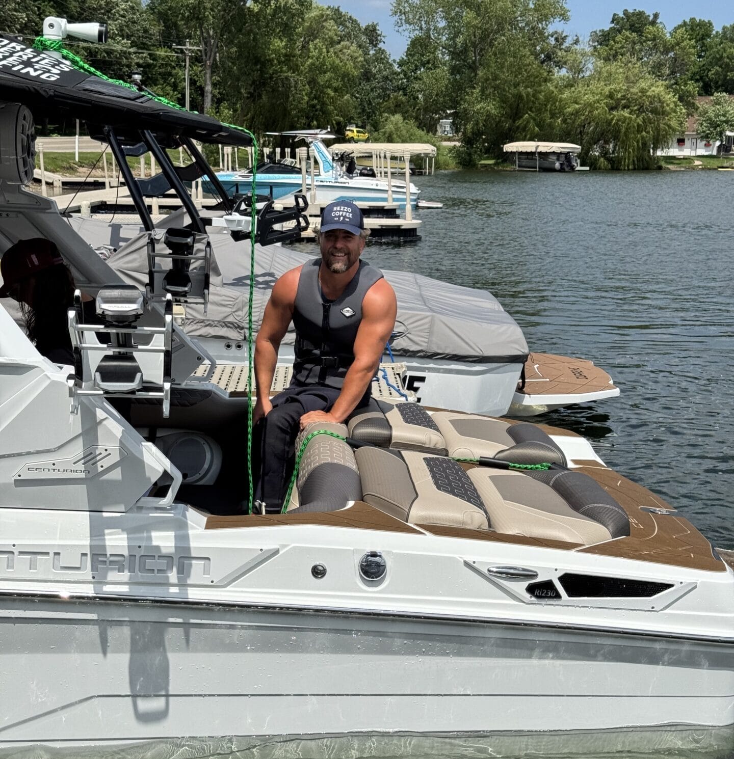 A person wearing a life jacket and cap sits on the edge of a boat docked on a lake, surrounded by trees and other boats.