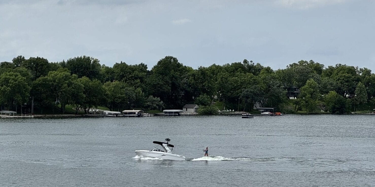A person is wakeboarding behind a white boat on a lake, with another boat nearby and trees lining the shore under a cloudy sky.
