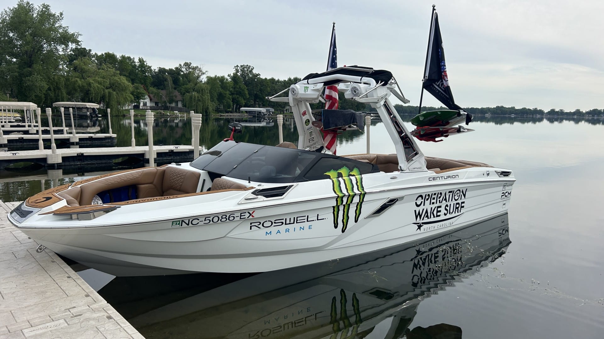A white Centurion boat with "Operation Wake Surf" and sponsor logos docked on a calm lake, with trees and docks in the background.