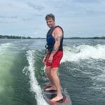A man wearing a life vest and red shorts is wake surfing on a lake under a partly cloudy sky.