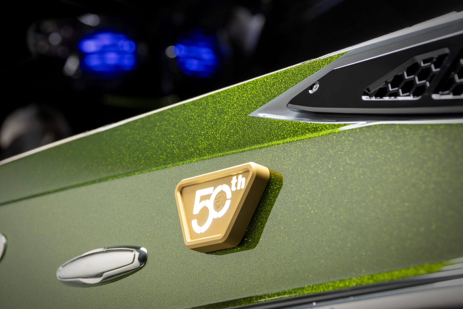 Close-up of a green metallic car with a gold "50th Anniversary" badge on the side, featuring chrome details and black vent trim.