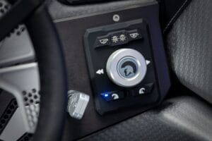 Close-up of a 2026 Ri245 vehicle control panel with illuminated buttons and a central dial, featuring icons for windshield, lighting functions, and optimized controls for water sports or boating activities.