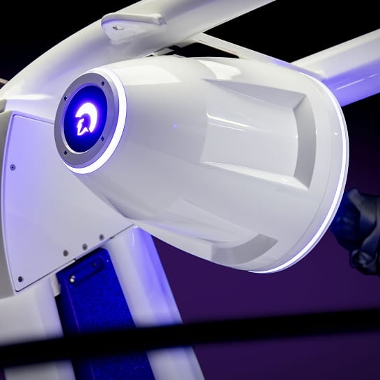 Close-up of a white, futuristic, cylindrical machine with blue LED lighting, mounted on a glossy structure against a dark purple background.