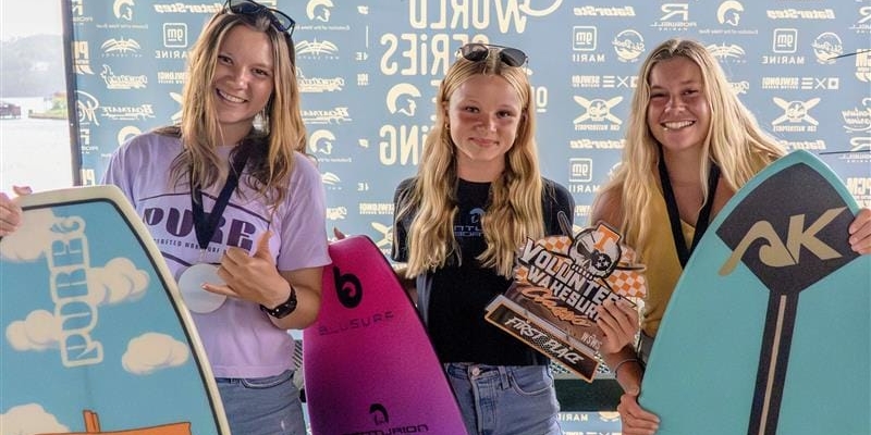 Three young women stand holding skimboards and a trophy in front of a branded backdrop at the 2025 Centurion WSWS Volunteer Wake Surf Classic Finals.