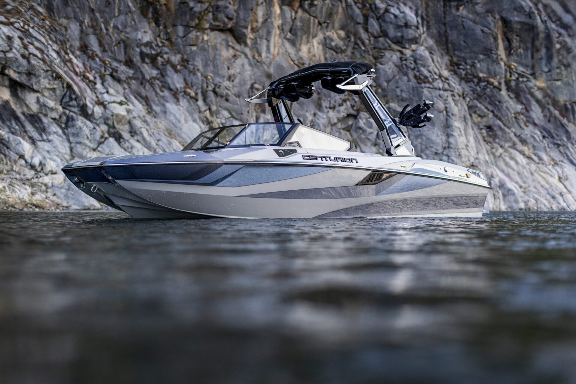 A modern 2026 Fe23 Centurion speedboat, a fearlessly evolved towboat, floats on calm water near a rocky cliff.