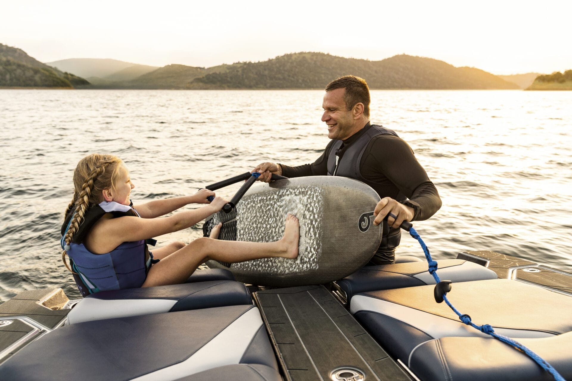 A man and a young girl wearing life vests sit on a boat at sunset, smiling as they hold a wakeboard between them—showing that teamwork is what Pass The Handle is all about.