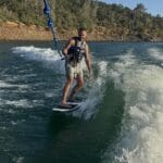 A man wearing a life vest and shorts rides a Centurion Boats wakesurf board while holding a blue rope on a lake, with the forested shoreline in the background.
