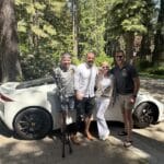 Four people stand and smile in front of a white convertible sports car parked on a shaded driveway with trees in the background. One person, supported by Ranger Road, has a prosthetic leg and uses crutches.