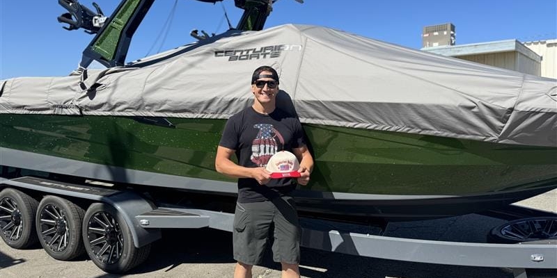 A person stands in front of a large covered Centurion Boat on a trailer, holding a small circular award. The scene is outdoors on a sunny day at a Ranger Road non-profit event.
