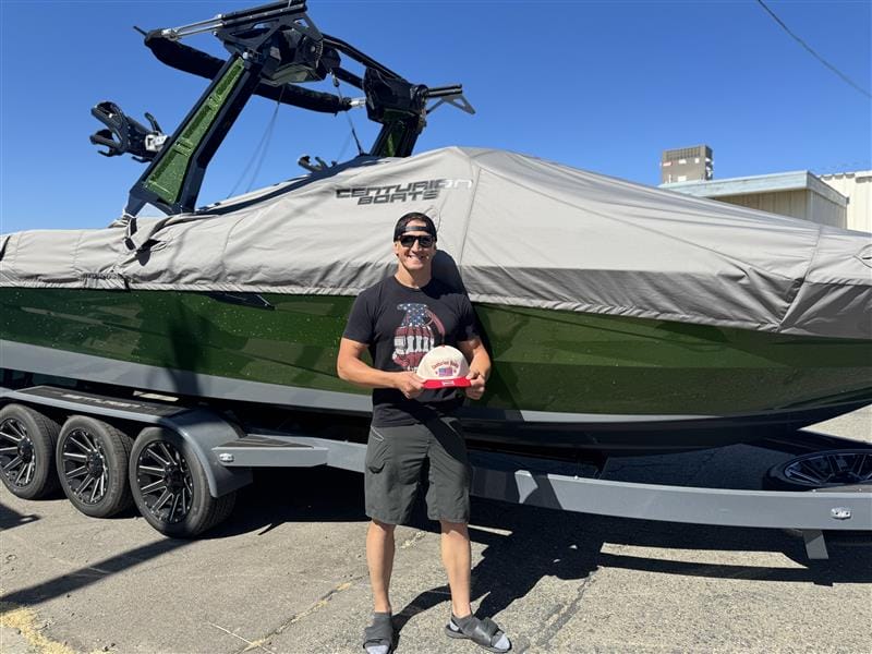 A person stands in front of a large covered Centurion Boat on a trailer, holding a small circular award. The scene is outdoors on a sunny day at a Ranger Road non-profit event.