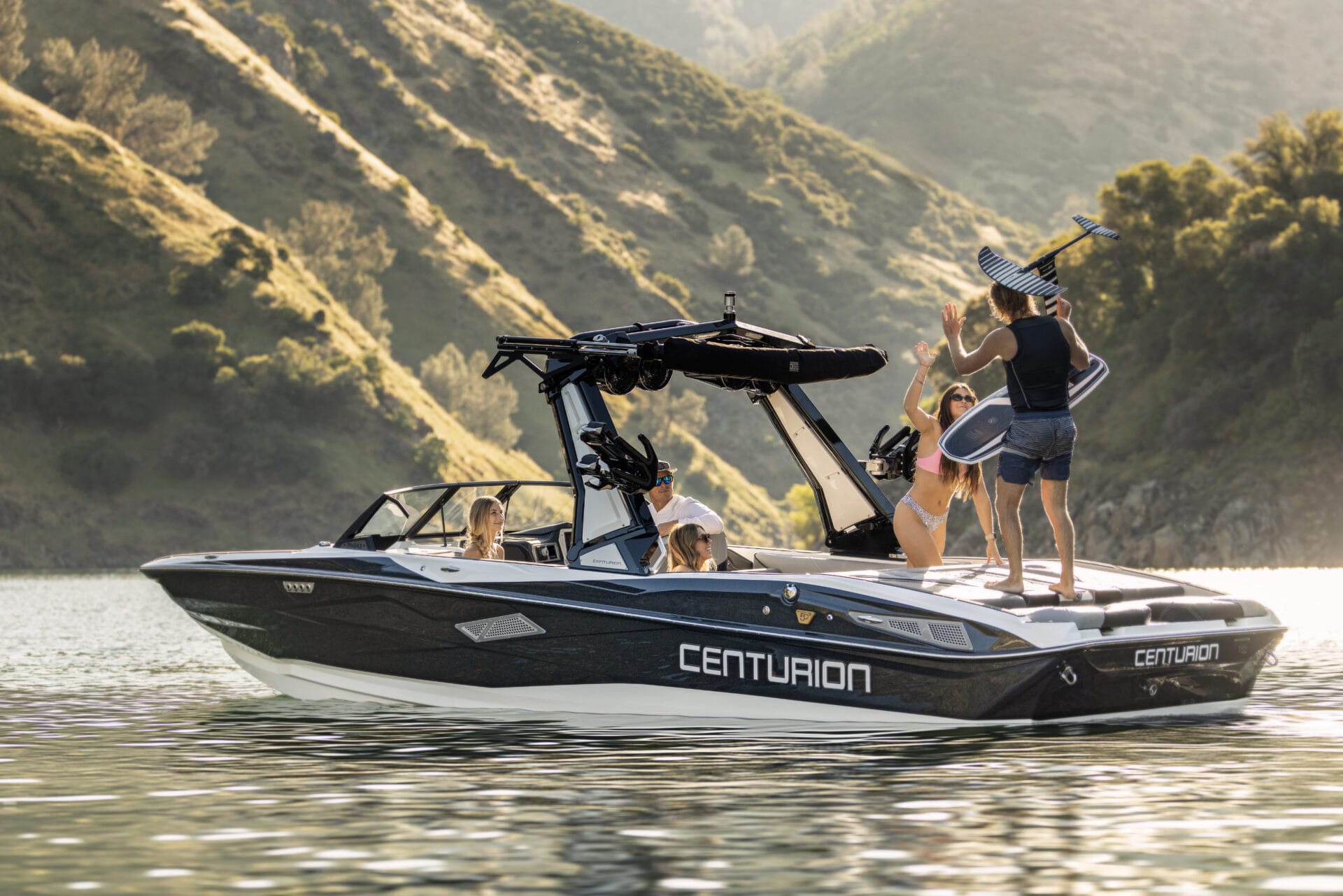 A group of people are on a 2026 Centurion motorboat, enjoying victory on the water near forested hills; one person proudly holds a wakeboard.