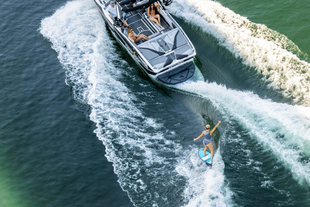 A person rides a wakeboard behind a motorboat on a lake, experiencing true Victory on the Water in style, while two others relax on the boat enjoying the scene.
