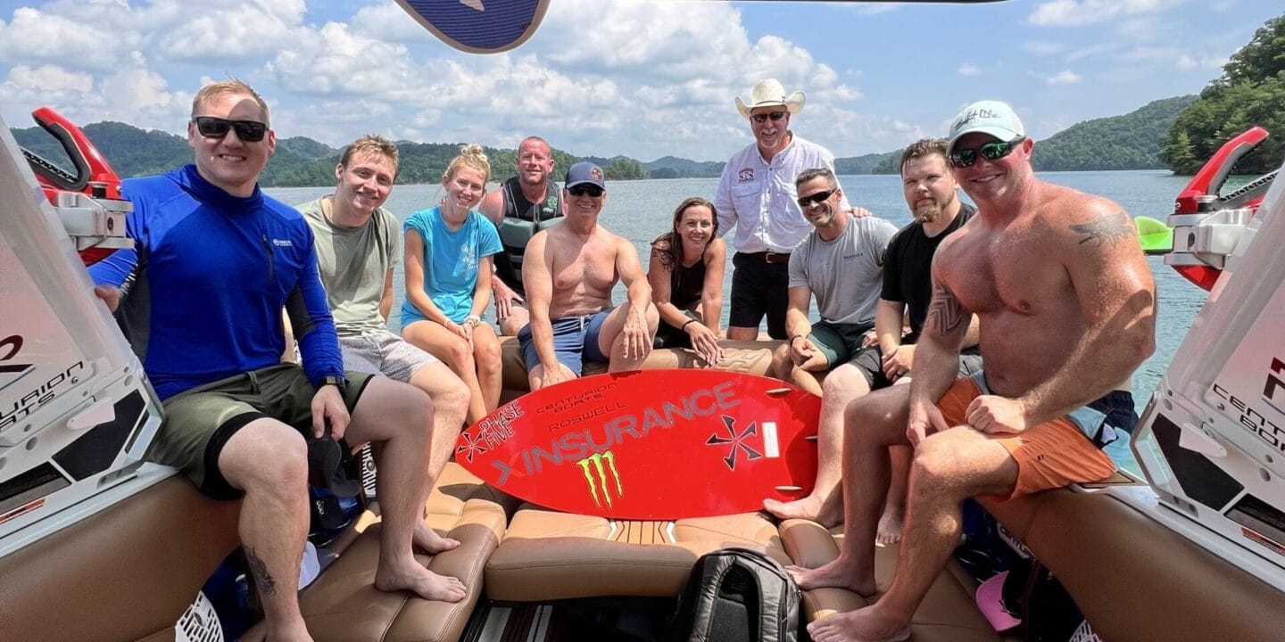 A group of people sit and pose for a photo on a Centurion Boat, some holding a red wakeboard during the Operation Wake Surf Honor Heroes Salute to Service Event on South Holston Lake, with trees and sky visible in the background.