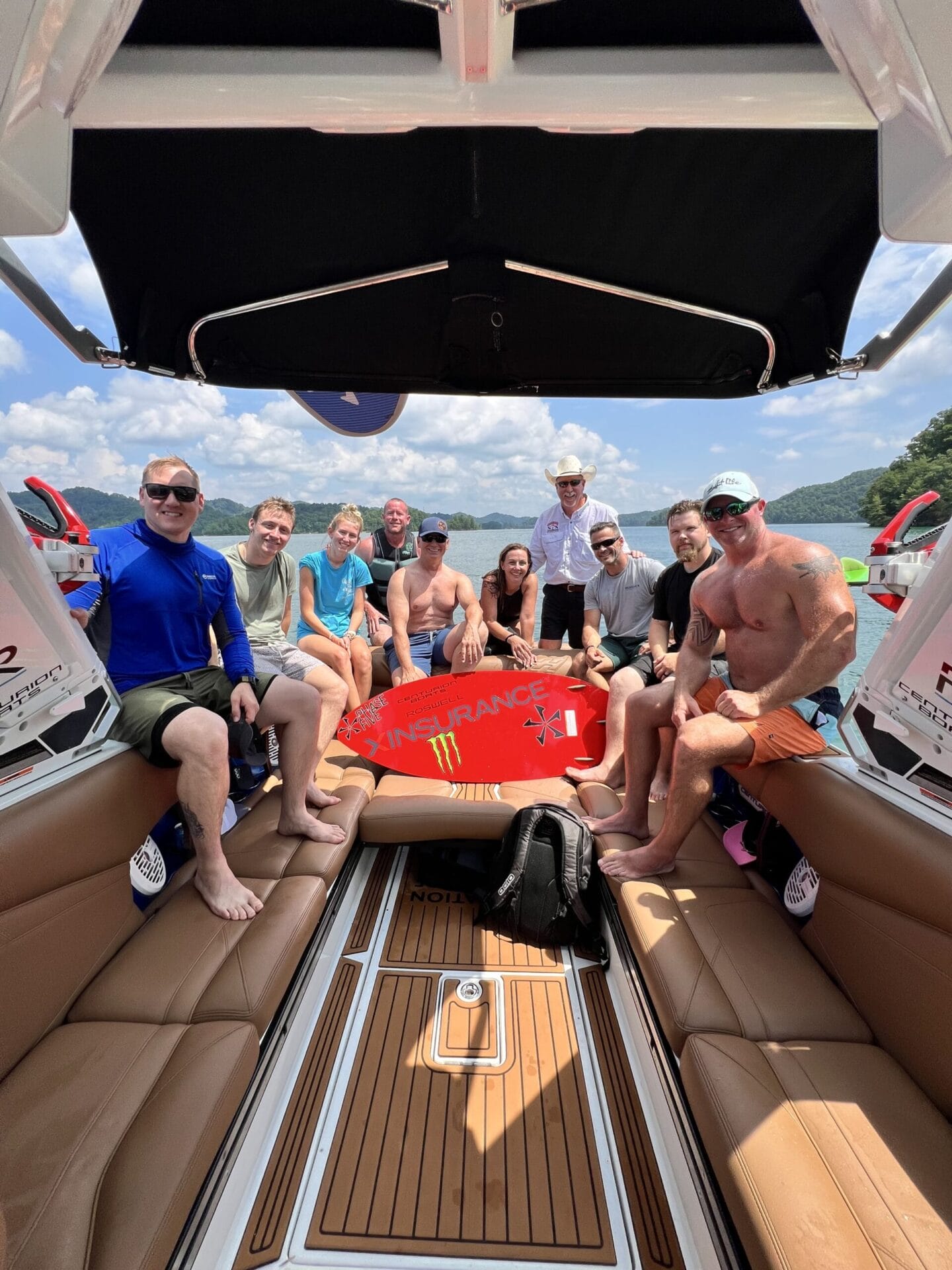A group of people sit and pose for a photo on a Centurion Boat, some holding a red wakeboard during the Operation Wake Surf Honor Heroes Salute to Service Event on South Holston Lake, with trees and sky visible in the background.