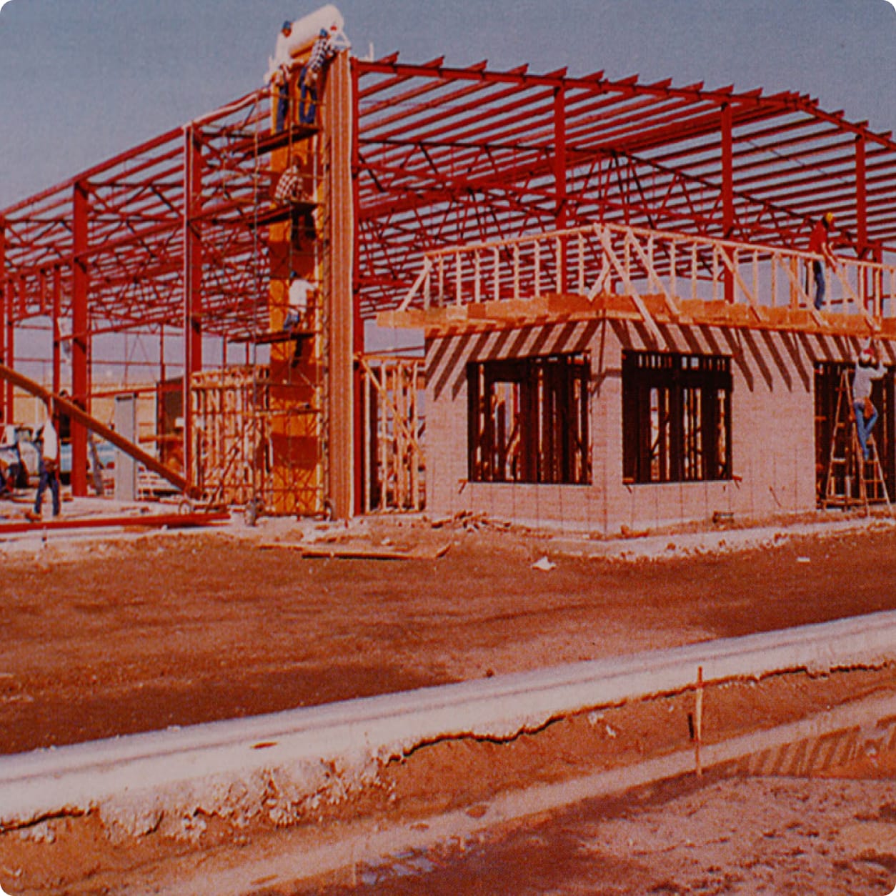 A building under construction with exposed steel framework, scaffolding, and workers on site resembles the sturdy design of a Centurion Opti-V Hull. The ground is bare with some puddles and construction materials visible.