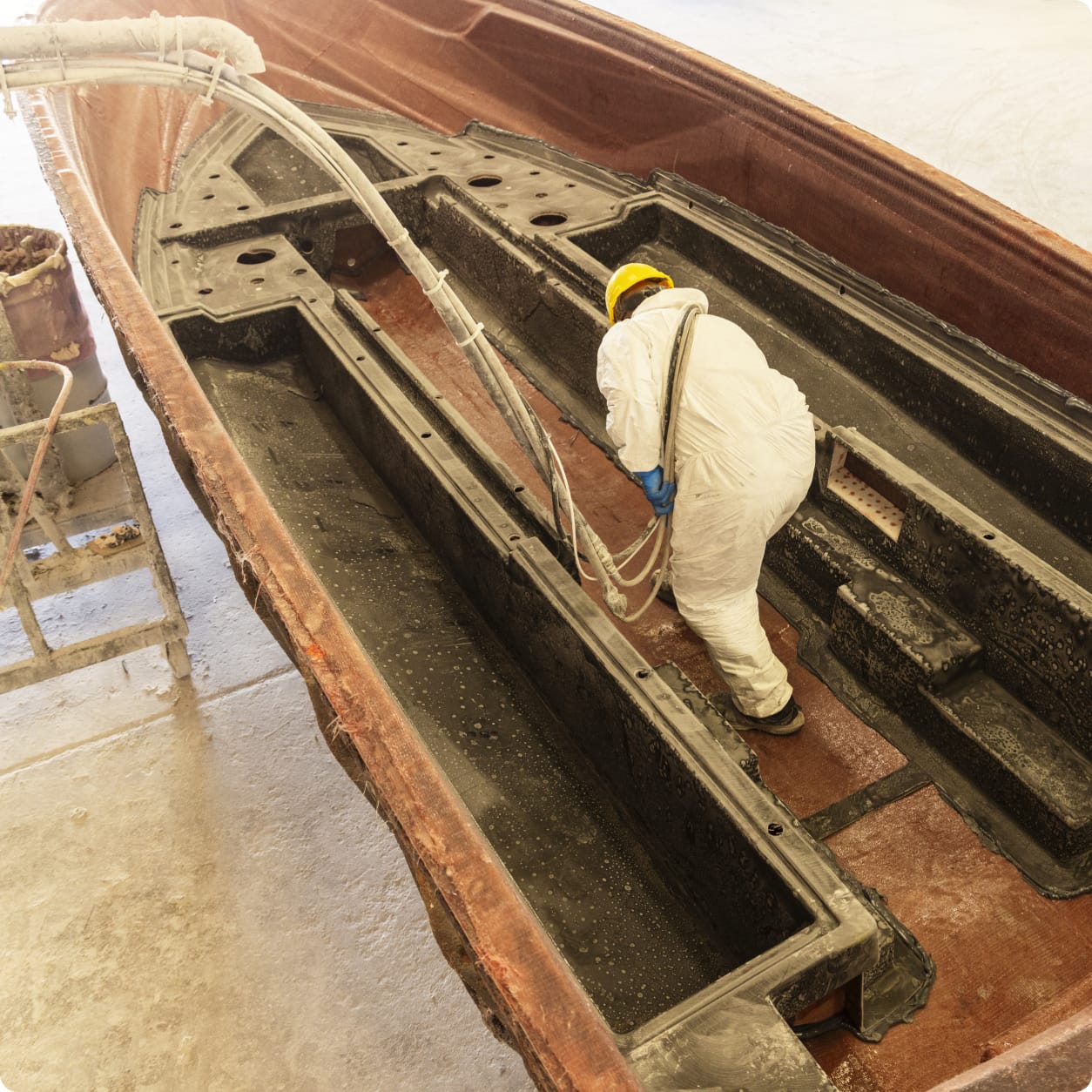 A worker in protective gear sprays insulation material inside a large, empty Centurion Ballast boat hull in an industrial setting.