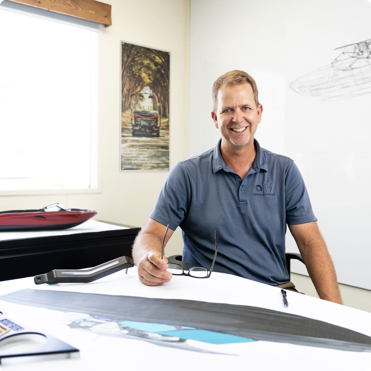 A man in a blue polo shirt sits at a desk with Centurion Opti-V Hull boat design drawings, holding glasses and smiling at the camera in a bright, modern office.