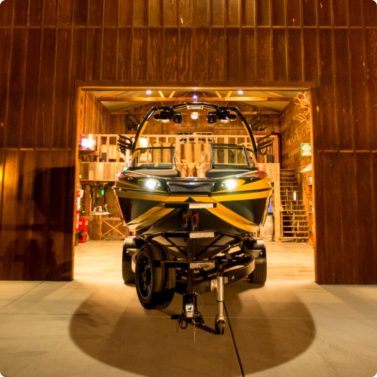 A Centurion Opti-V Hull speedboat on a trailer with headlights on is parked inside a wooden barn with open doors at night.