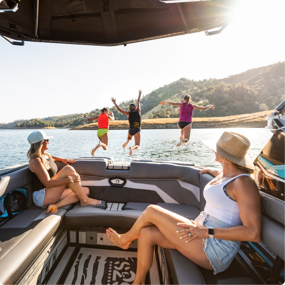 Four people leap into a lake from a boat featuring a Centurion Vinyl interior, while two others relax onboard, surrounded by scenic hills and trees in the background.